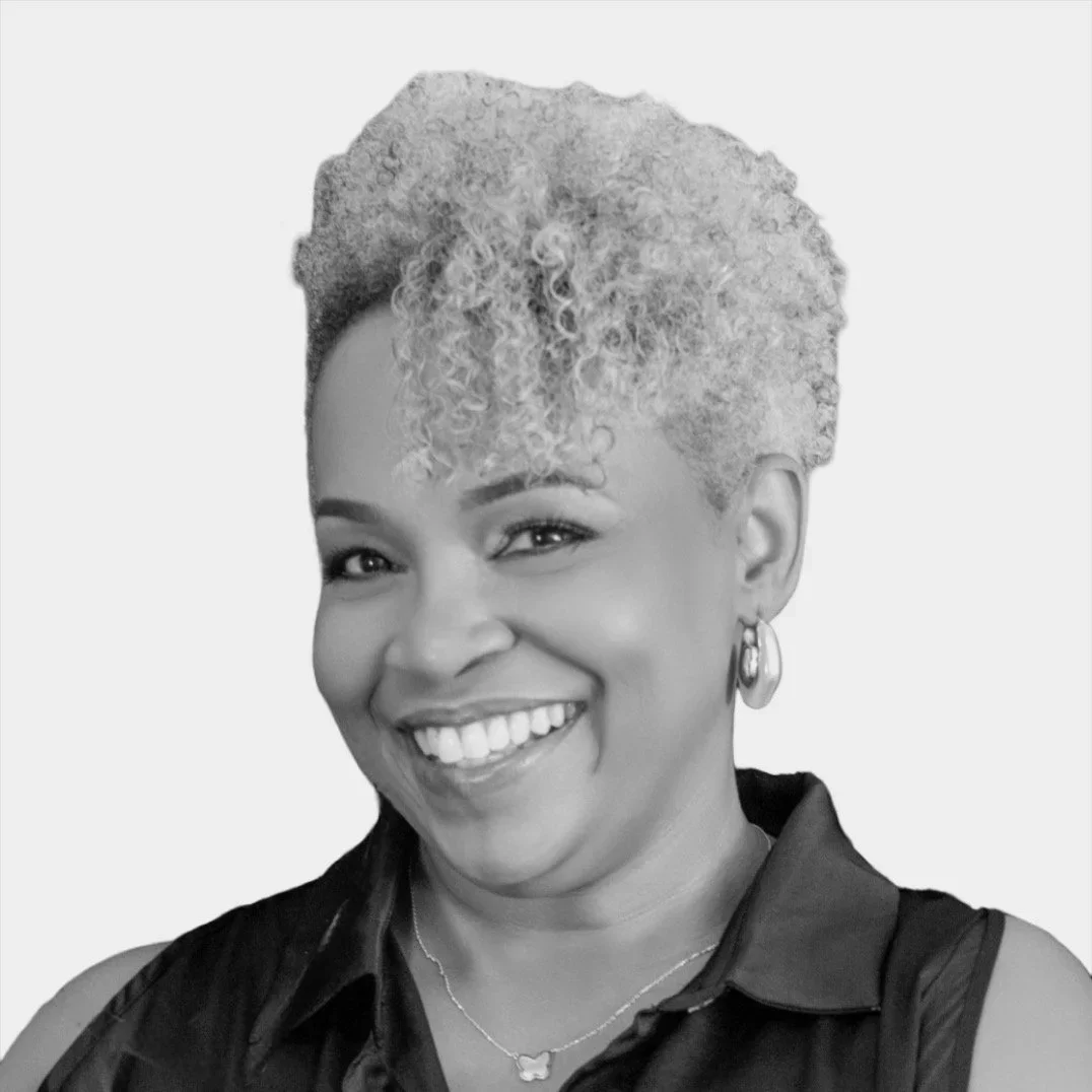 Black and white portrait of a joyful woman with short, curly hair, wearing earrings, a necklace with a butterfly pendant, and a sleeveless top.