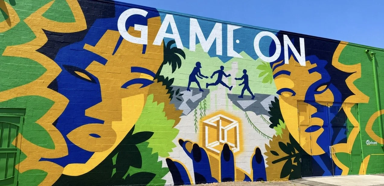 Colorful mural with the words "GAM ON" at the top, depicting silhouettes of people playing, walking, and holding hands, surrounded by abstract shapes and greenery.