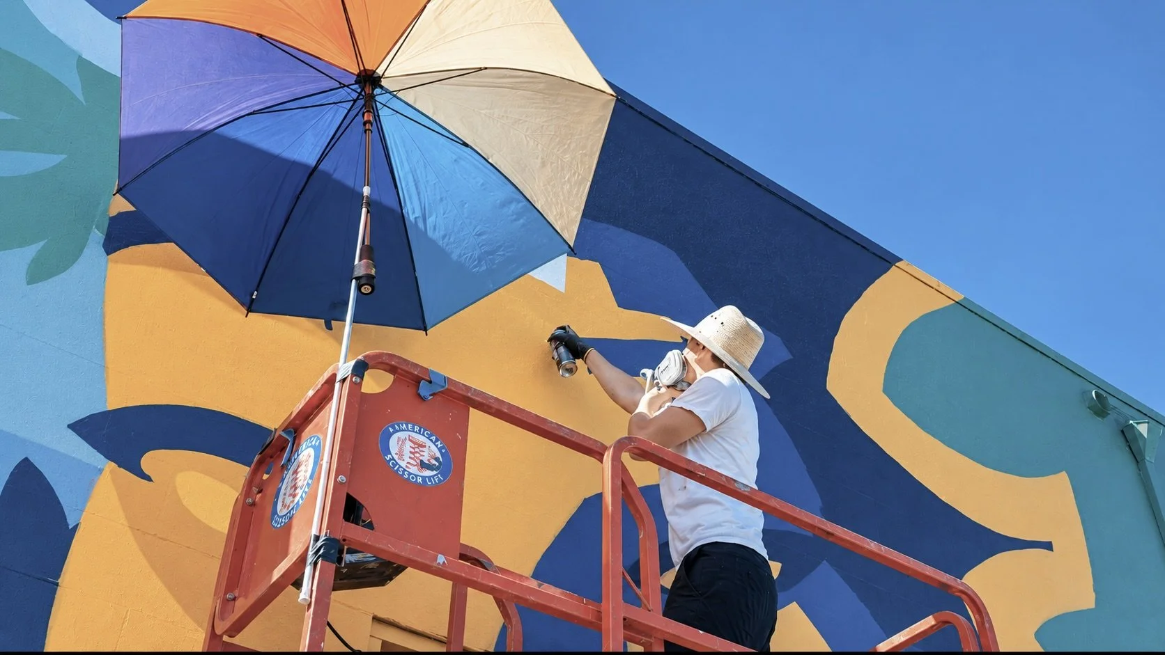 A person standing on a cherry picker wearing a straw hat, mask, gloves, and white t-shirt, painting a large mural on a building exterior under a blue sky with an umbrella providing shade.