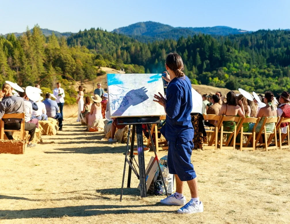 An outdoor art event, with a young artist painting on an easel while a crowd watches in front of a scenic mountain and forest backdrop.