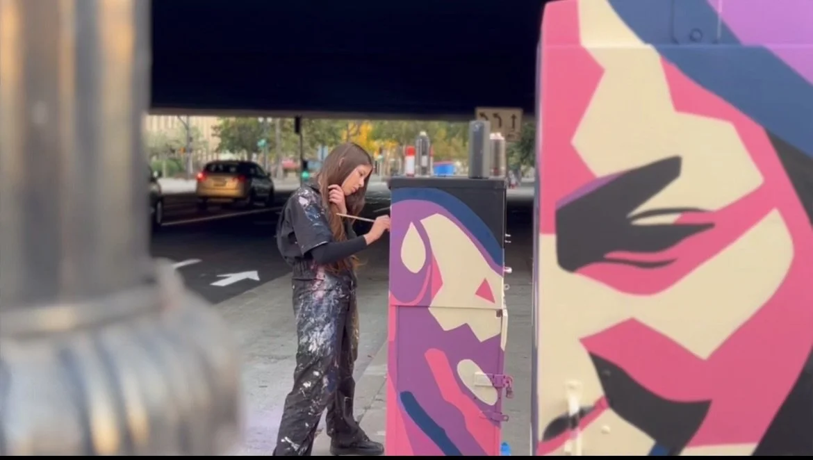 A woman painting graffiti art on a utility box on a city sidewalk, with a street and cars visible in the background.