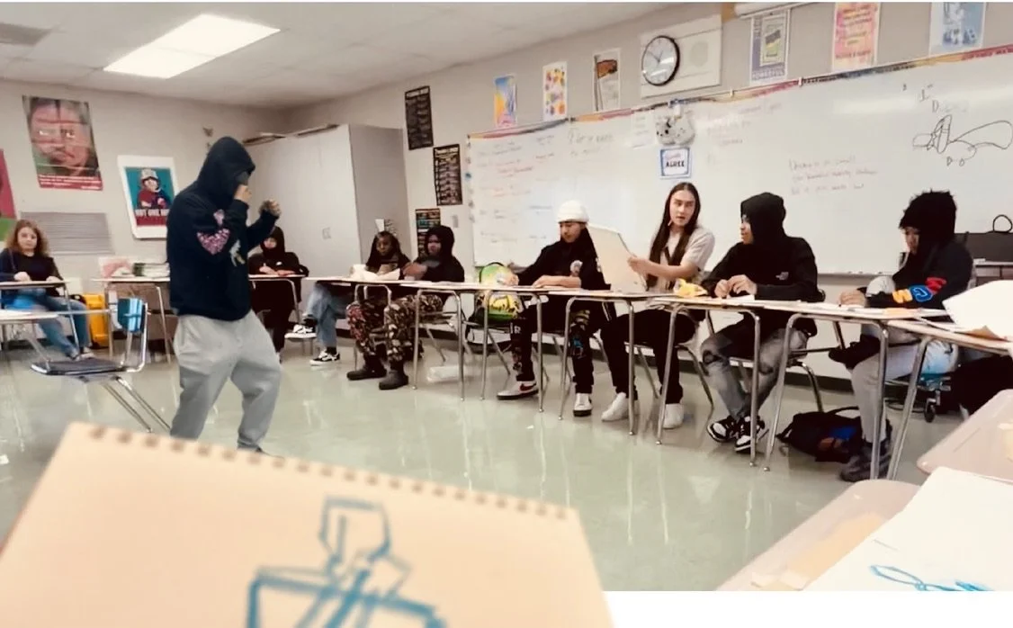 A classroom with students seated at desks and a person standing in the center, possibly presenting. The whiteboard has writing and doodles, and posters are on the walls.