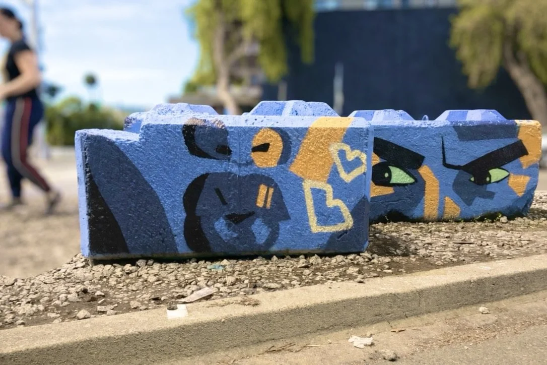 Graffiti art on three blue concrete blocks, featuring a lion's face, a cat's eyes, and yellow hearts with blue outlines, outdoors on a gravel surface.