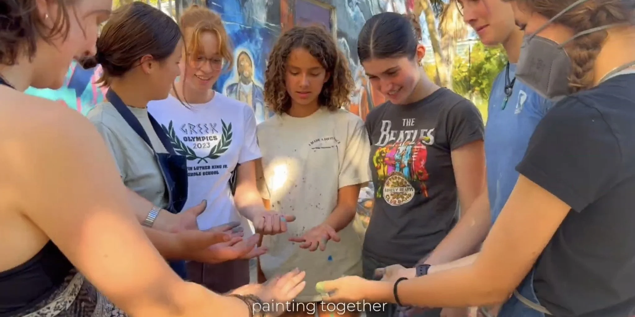 A group of young women are standing in a circle outdoors, painting their palms with colorful paint. They appear to be engaged in a shared creative activity, smiling and focused on their hands. The background shows a vibrant mural on a wall.