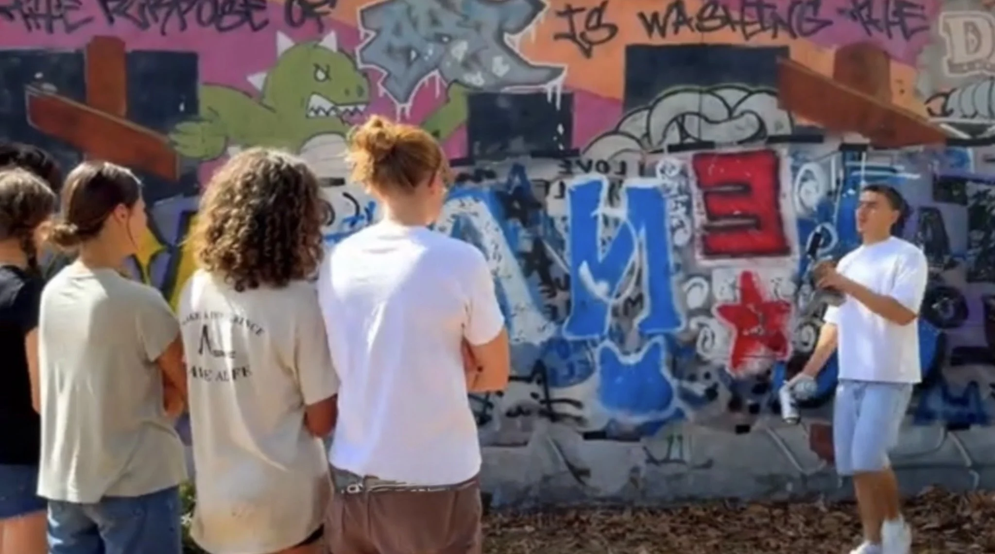 A group of young people stands near a colorful graffiti-covered wall. The wall features various designs, including a green dinosaur, words, and abstract art. One person appears to be talking while others listen.