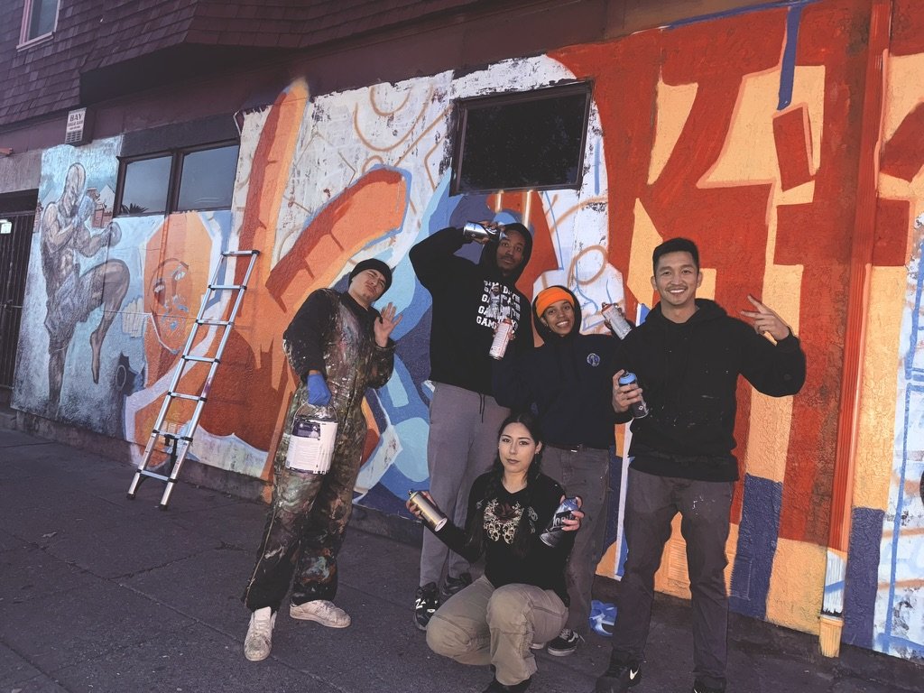 Group of five people posing with spray paint cans in front of a colorful mural on a city wall, with one person kneeling and the others standing, some making peace signs, a ladder leaning against the wall, and a black TV screen above the mural.