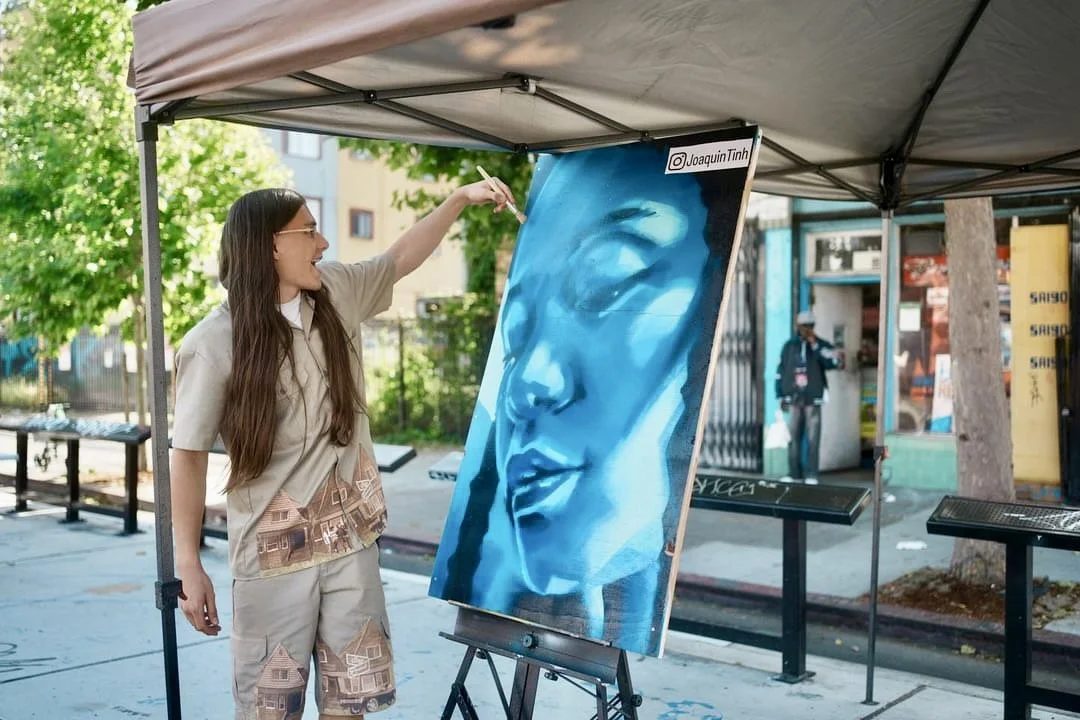 A woman painting a portrait of a woman's face on a large canvas under a tent outdoors.
