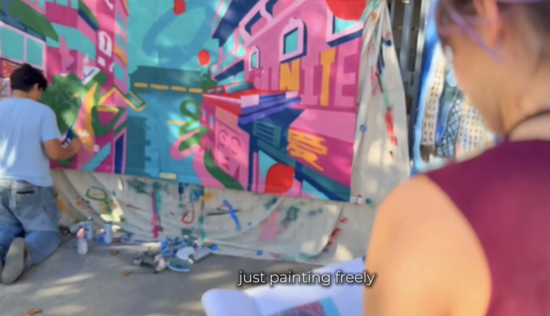 People painting a large, colorful mural on a wall outside. The mural features abstract shapes and vibrant colors like pink, blue, and green.