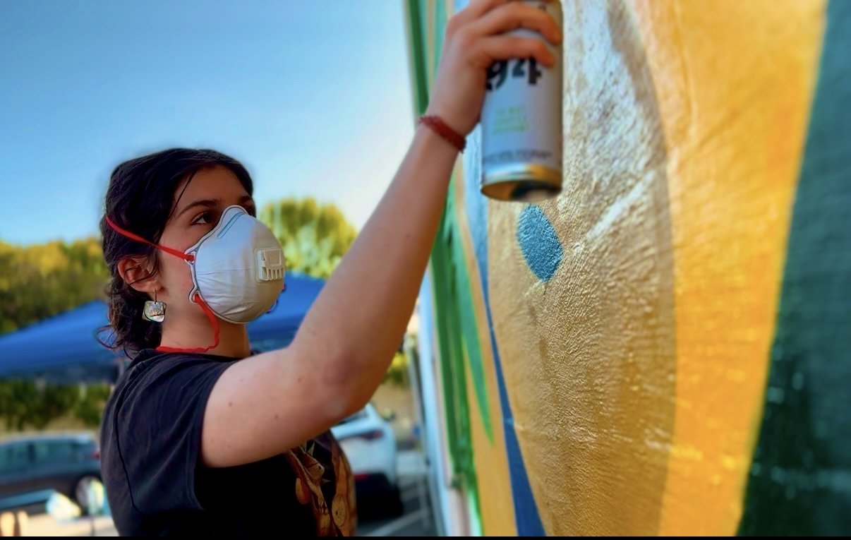 A woman wearing a face mask spray-painting a colorful mural outdoors.