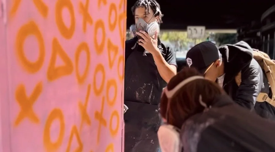 Three people wearing masks crowd around a colorful pink wall, with a woman partially hidden behind it, studying the wall or an artwork on it.