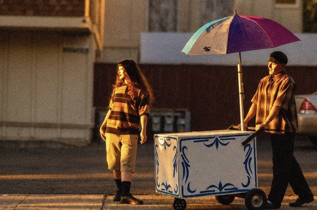 A young woman standing on the street at sunset, facing a young man behind a colorful ice cream cart with a large umbrella, smiling and engaged in conversation.