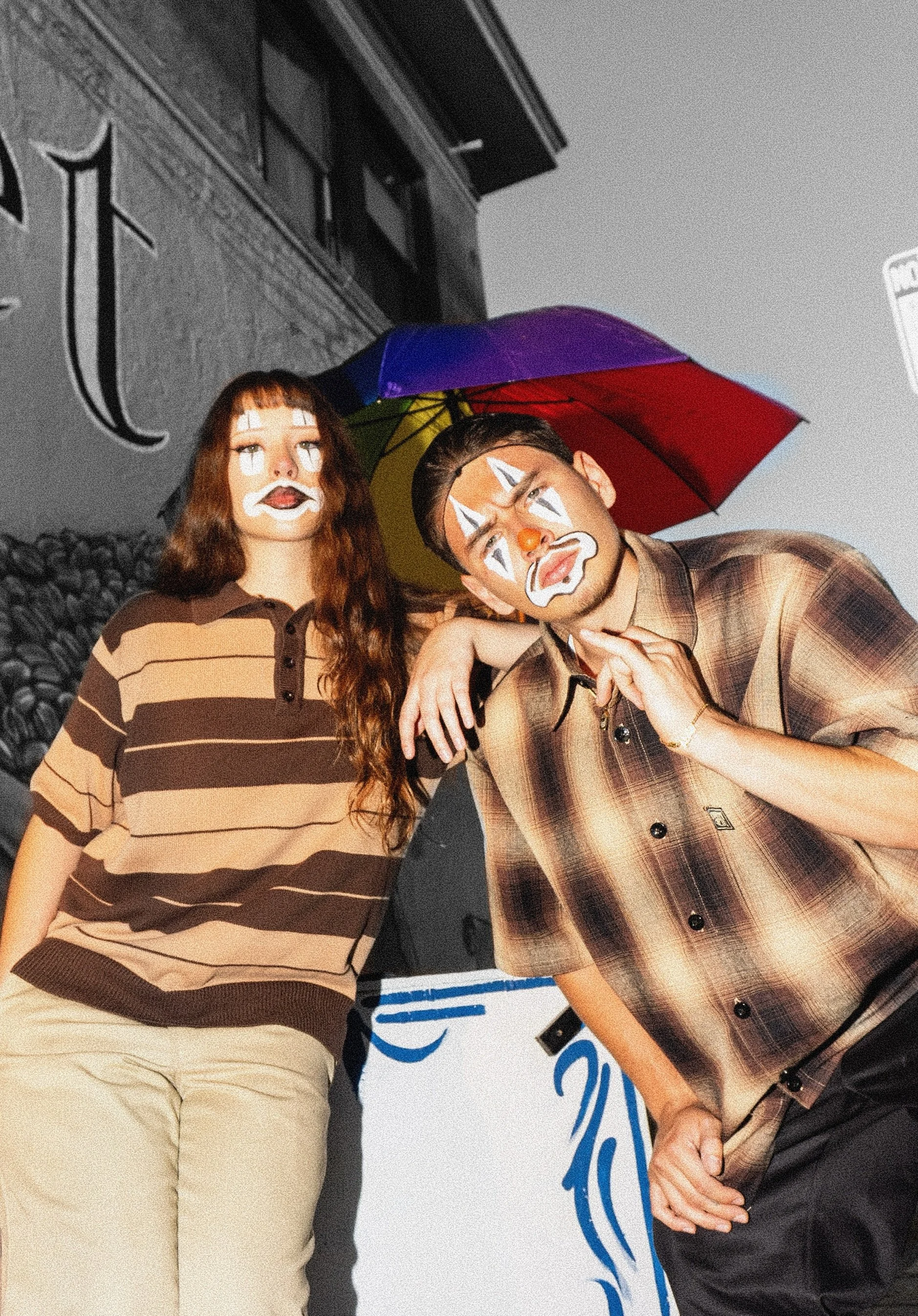 Two people dressed as clowns, holding an umbrella, standing next to each other at an event with a white and blue backdrop and some buildings behind