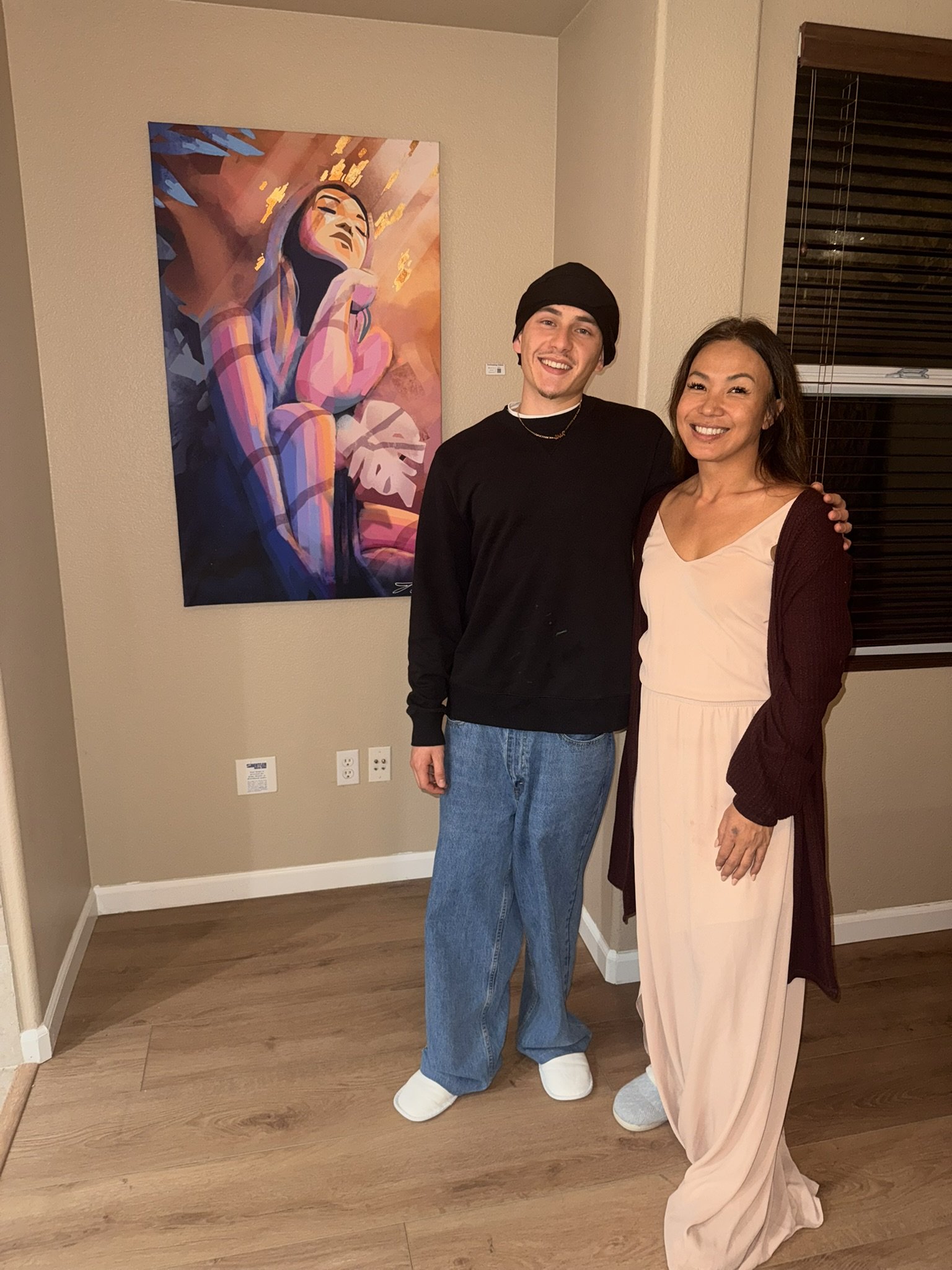 Two people standing next to each other indoors near a wall with a colorful portrait. They are smiling and wearing casual clothing, with one person in a black sweatshirt and jeans, and the other in a light dress with a dark cardigan. Both are wearing slippers.