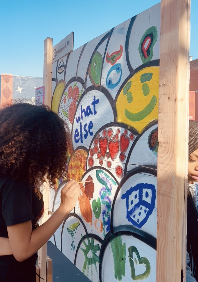 A woman with curly hair painting a mural on a wooden panel outdoor during daytime. The mural features colorful circles with various drawings, including a smiley face, hearts, and abstract art, with the words 'what else' written in blue.