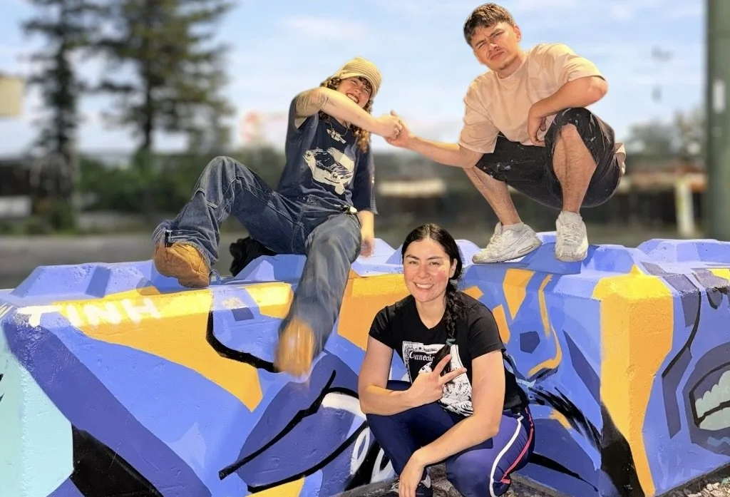 Three young people posing on a colorful graffiti-covered concrete structure outdoors. One person is sitting and the other two are squatting, all smiling and making playful gestures.
