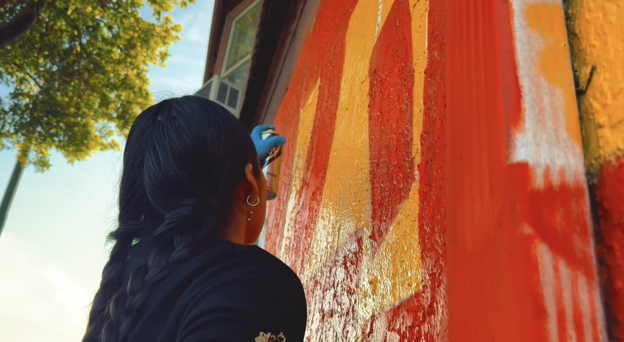 A person with long dark hair in a braid, wearing earrings and a black shirt, painting a colorful mural on a wooden surface outdoors. The mural features stripes in red, orange, and yellow. There is a tree and a house in the background, with blue sky and clouds.