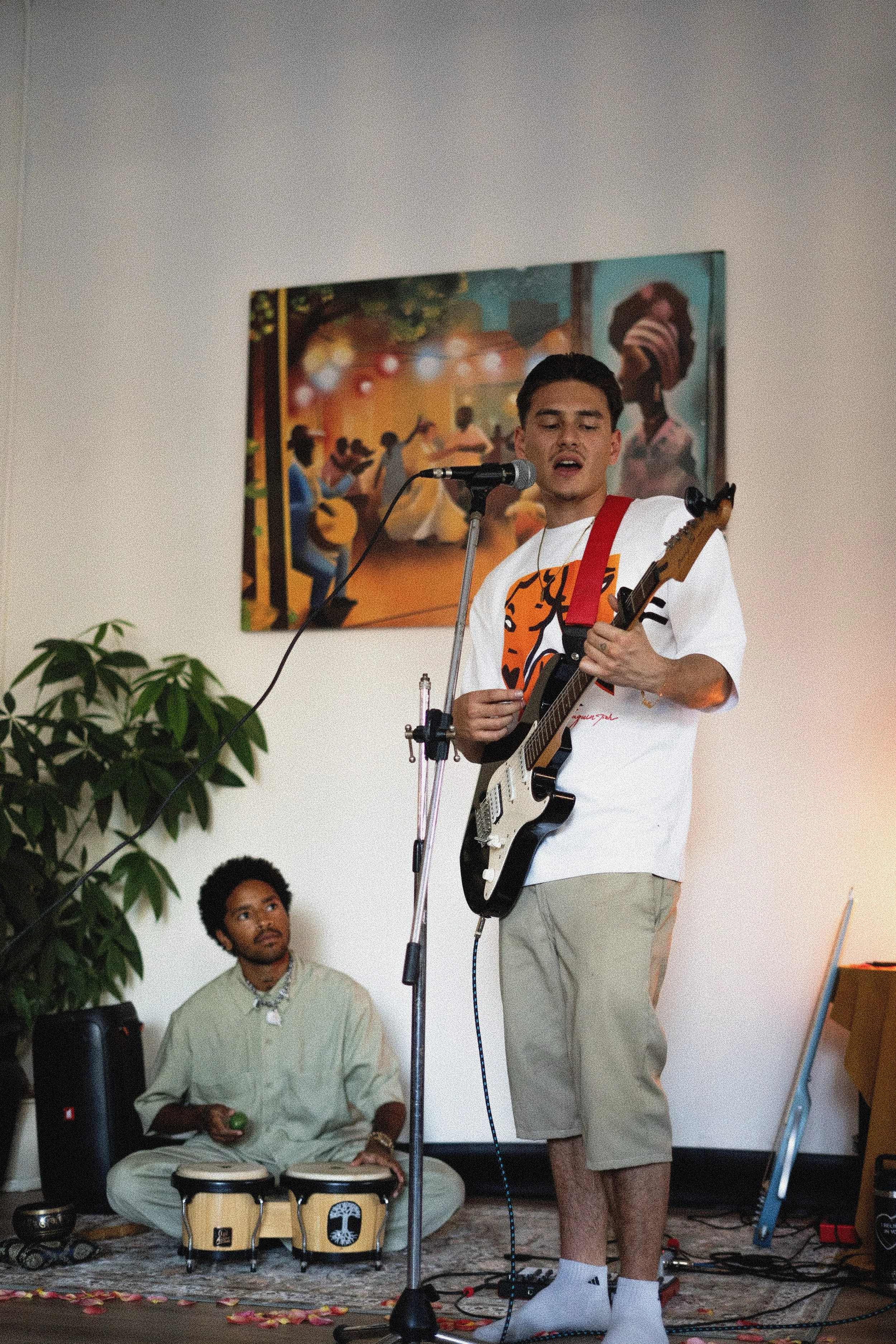 A young man with short dark hair singing and playing an electric guitar on a stage, wearing a white t-shirt with an orange graphic and khaki shorts. A man with afro hairstyle in beige clothing and wristbands plays bongos on the floor next to him. A woman with a headwrap and glasses is on a painting on the wall in the background. The stage is decorated with scattered flower petals, a blue guitar leaning against the wall, and a green plant in the foreground.