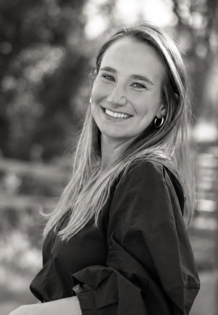 Black and white photo of a smiling woman with long hair, wearing hoop earrings and a dark top, outdoors with blurred trees in the background.