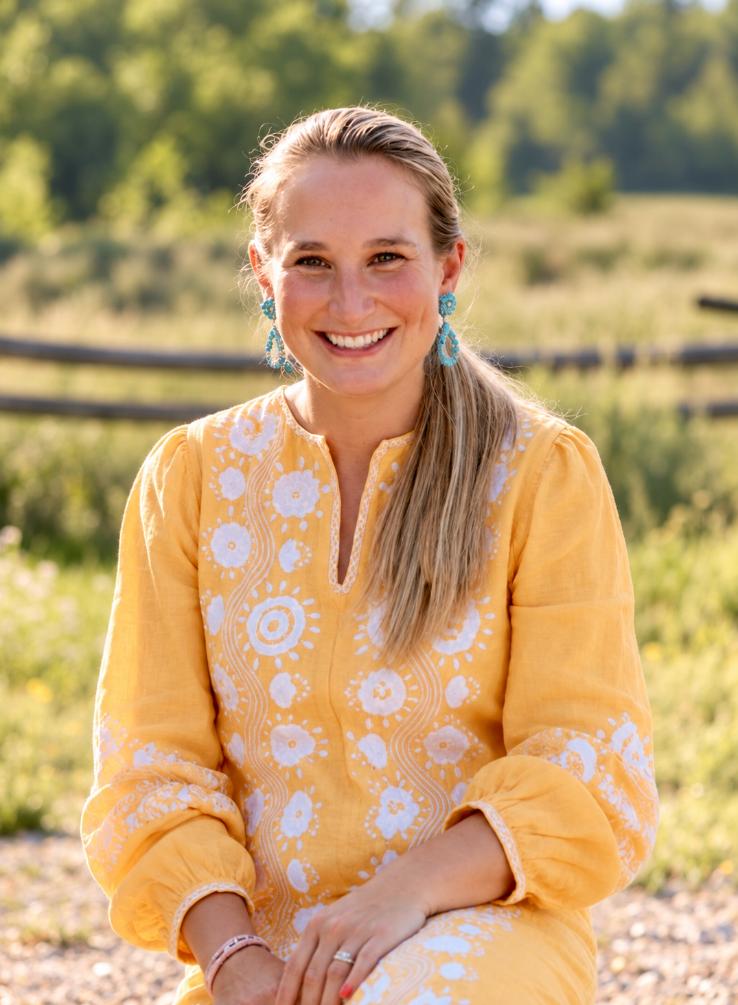 Smiling woman wearing a yellow embroidered dress and turquoise earrings outdoors in a sunny, green field.
