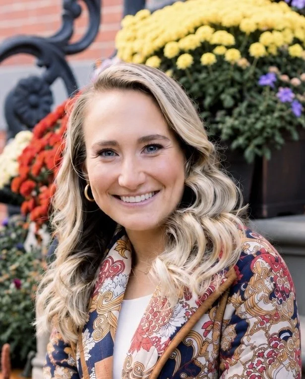 A smiling woman with blonde wavy hair, wearing a patterned blazer, standing outdoors near colorful flowers.