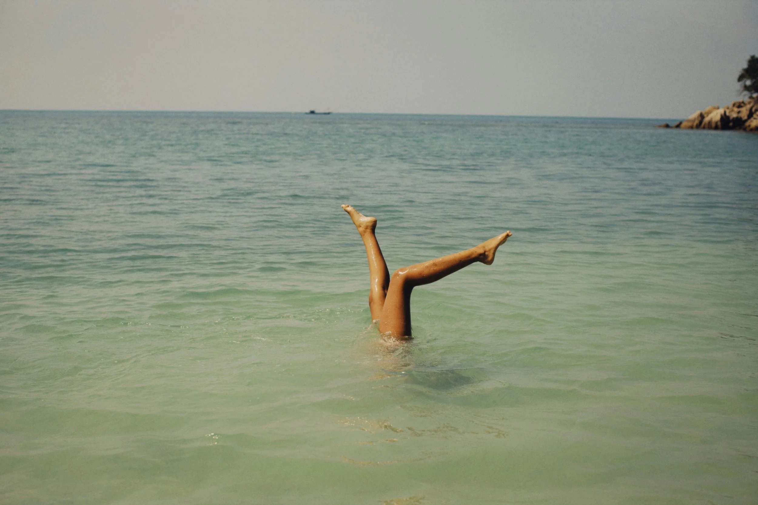 Person floating in the ocean with legs and feet sticking out of the water, with a rocky shoreline and a boat in the distance.