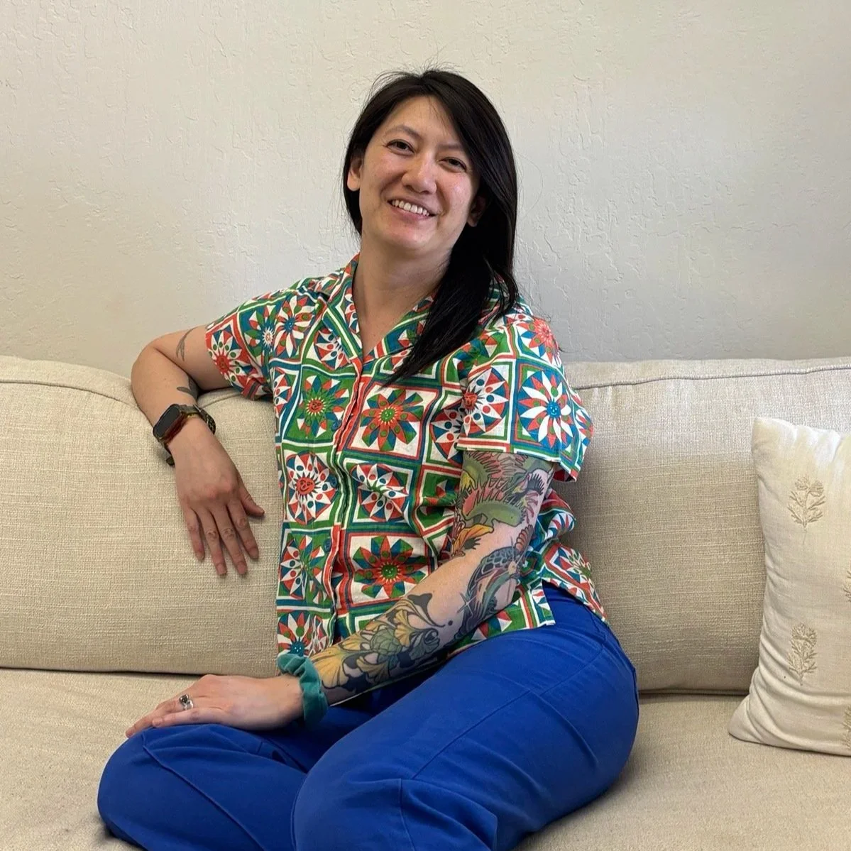 A female presenting person wearing a colorful short-sleeved, button-up shirt patterned with geometric shapes and bright blue pants smiles while casually sitting on a white couch.