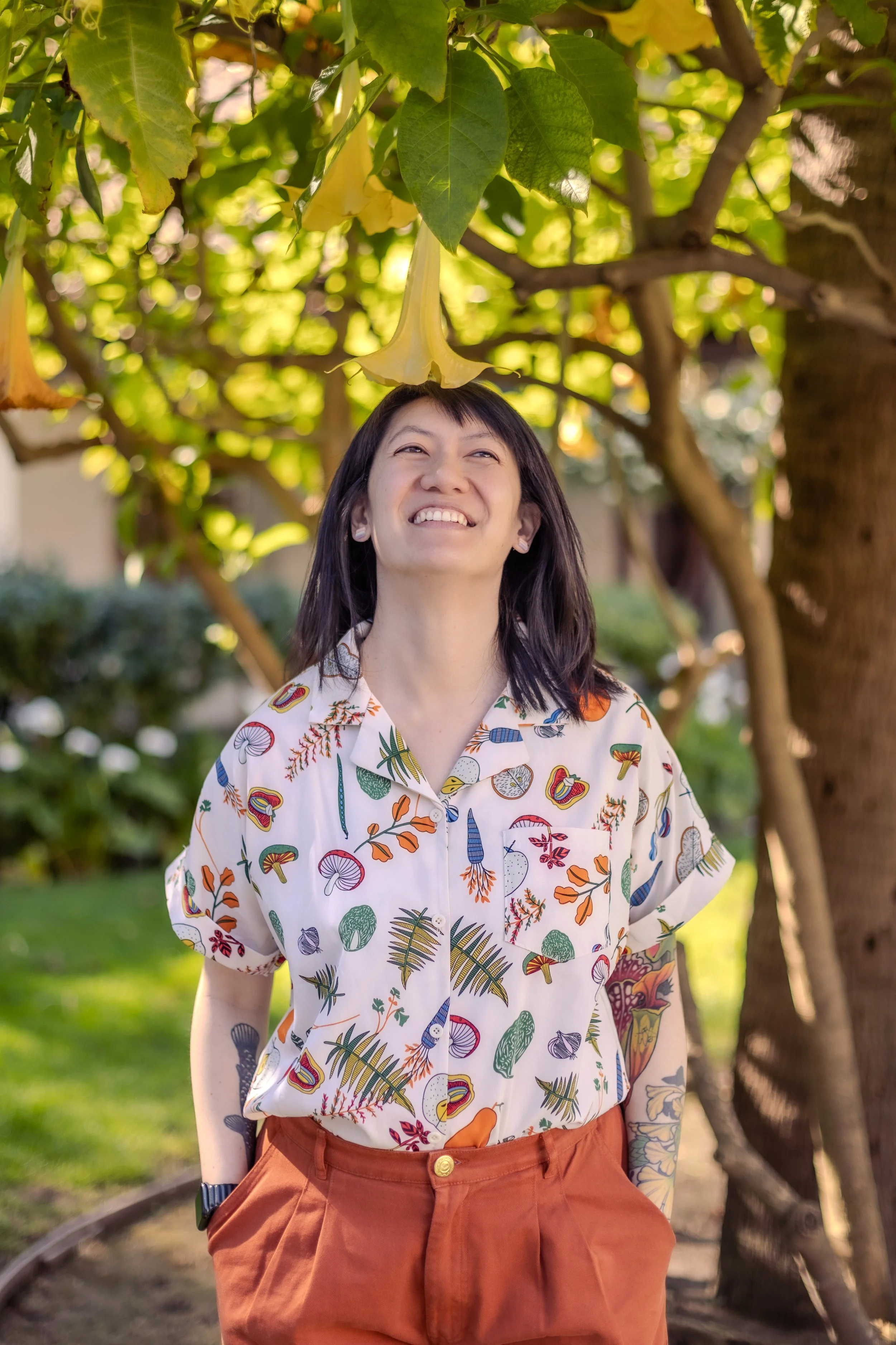 A tattooed female presenting person wearing a colorful short-sleeved, button-up shirt patterned with mushrooms and leaves smiles while standing under and looking up at a yellow flower descending from a tree branch.
