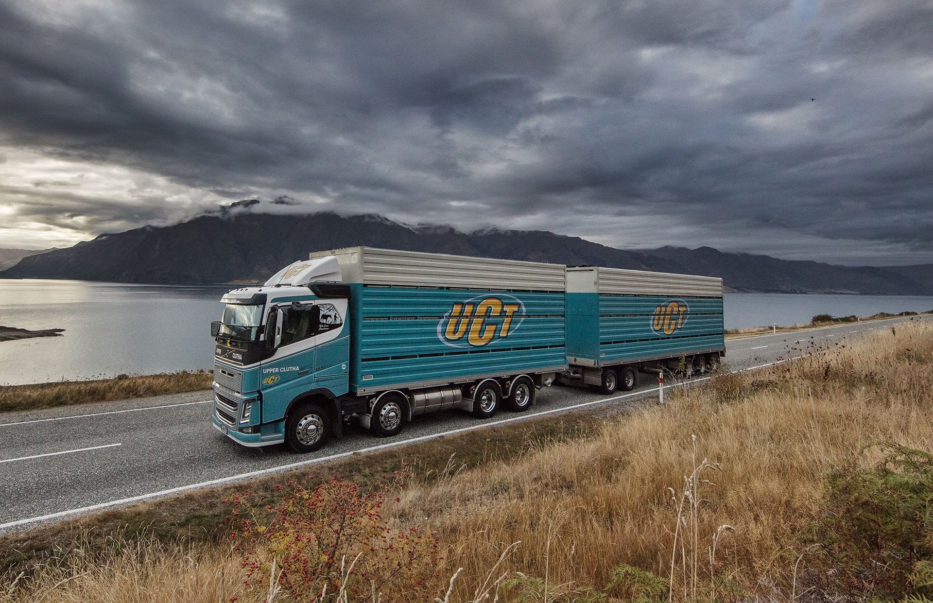 A large teal UCT semi-truck driving along a scenic highway beside a lake with mountains in the background on a cloudy day.