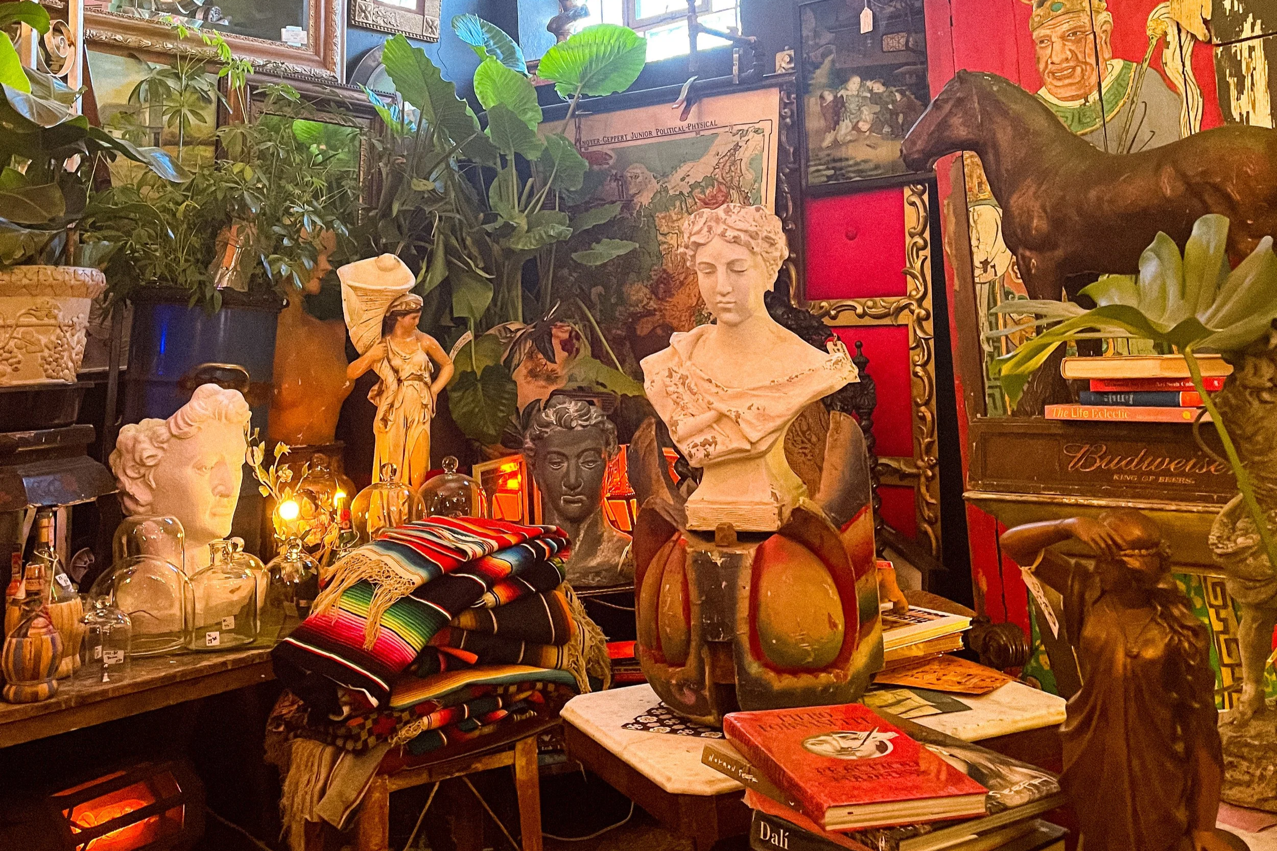 A room with vintage and eclectic decorations, including bust sculptures, potted plants, framed pictures, books, and colorful textiles. The space has a warm and artistic atmosphere.