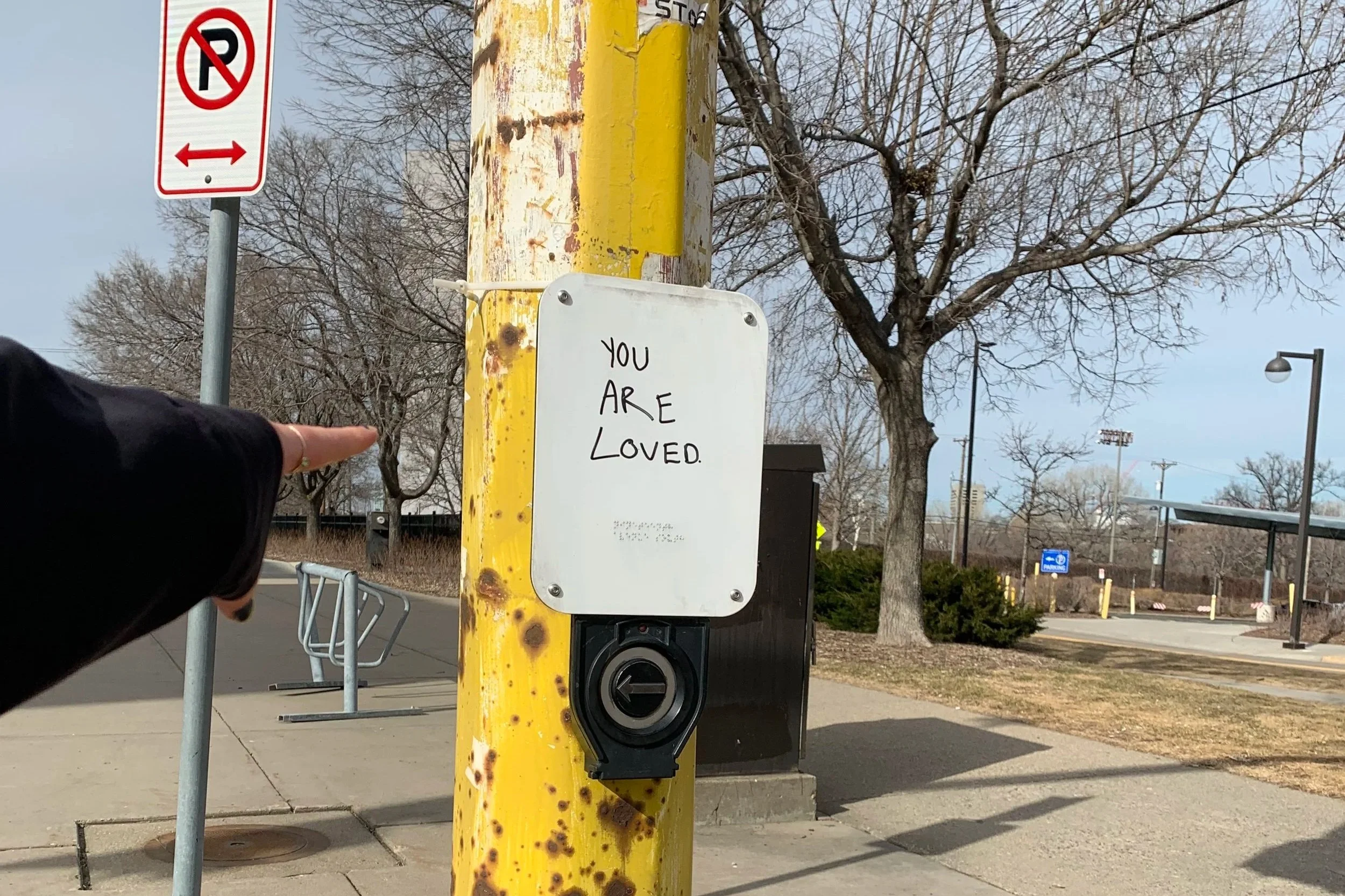 A white sign on a yellow metal post with the handwritten message "You Are Loved" in black marker. The background shows leafless trees, streetlights, and a parking lot.