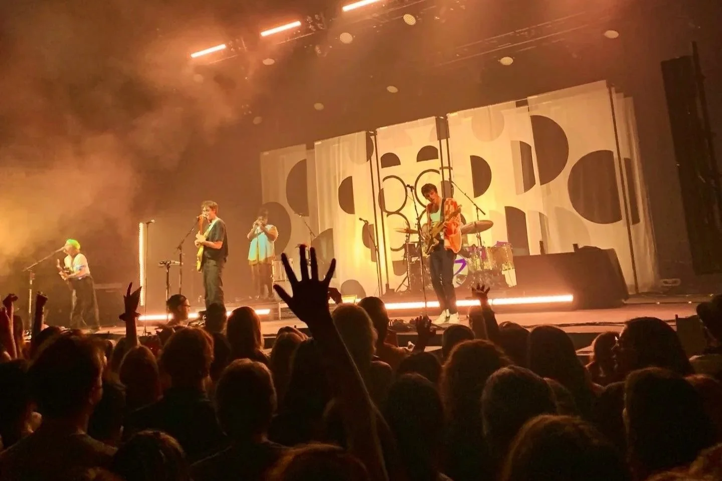 A live concert scene with a band performing on stage. The band consists of five members playing various instruments, including guitars and drums. Audience members are visible, some with hands raised, enjoying the performance. The stage features geome