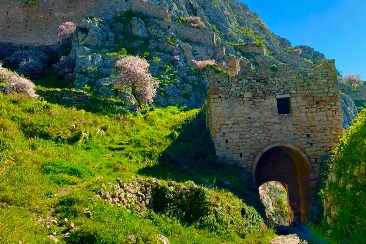 Scenic landscape with a stone archway and ancient stone building, blooming pink and purple trees, green grass, and rocky hillside under a blue sky.