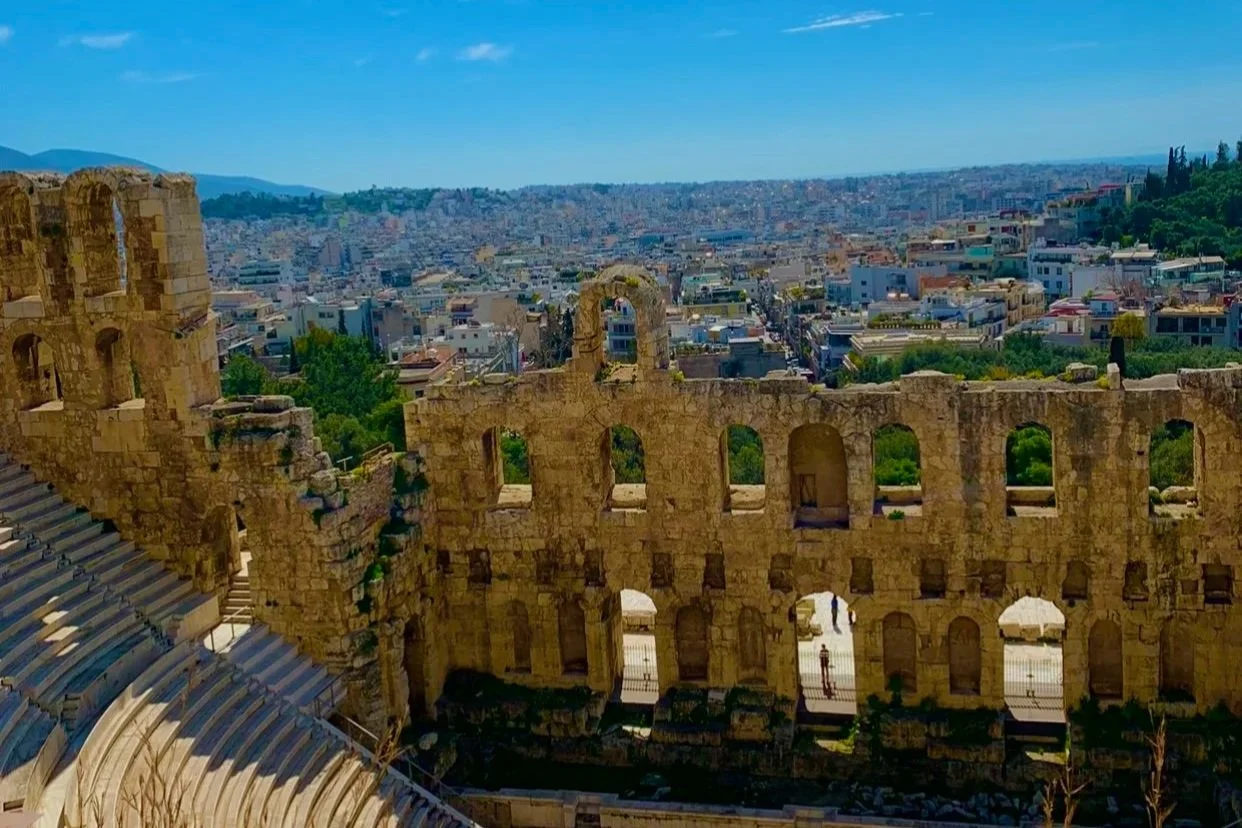 Ancient Greek or Roman amphitheater ruins with a cityscape in the background under a clear blue sky.