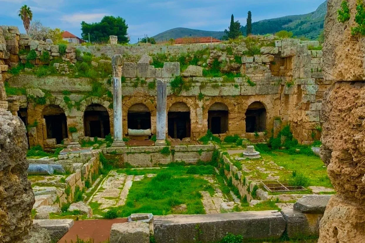 Ancient Roman ruins with stone walls, columns, and an open courtyard overgrown with grass and small plants.