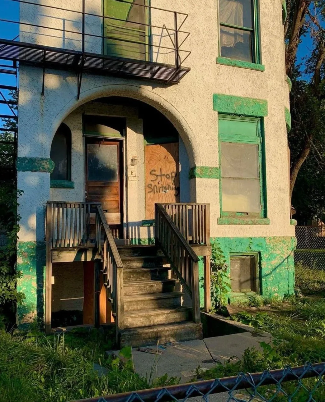 An old, abandoned house with peeling white and green paint, boarded-up front door, and broken windows. A wooden staircase leads to the entrance, with a sign on the door saying "Stop Snitching." The house appears neglected, with overgrown grass and pl