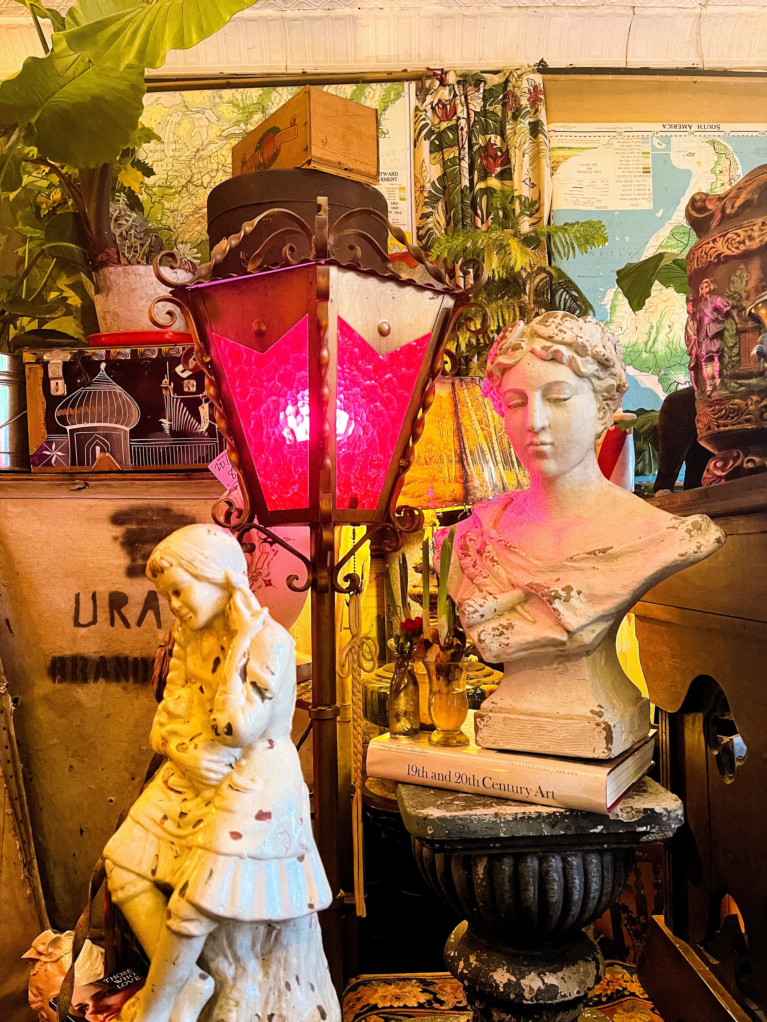 Interior of an antique shop with sculptures, lamps, and vintage decor, including a bust of a woman and a smaller sculpture of a girl, as well as plants, maps, and books.