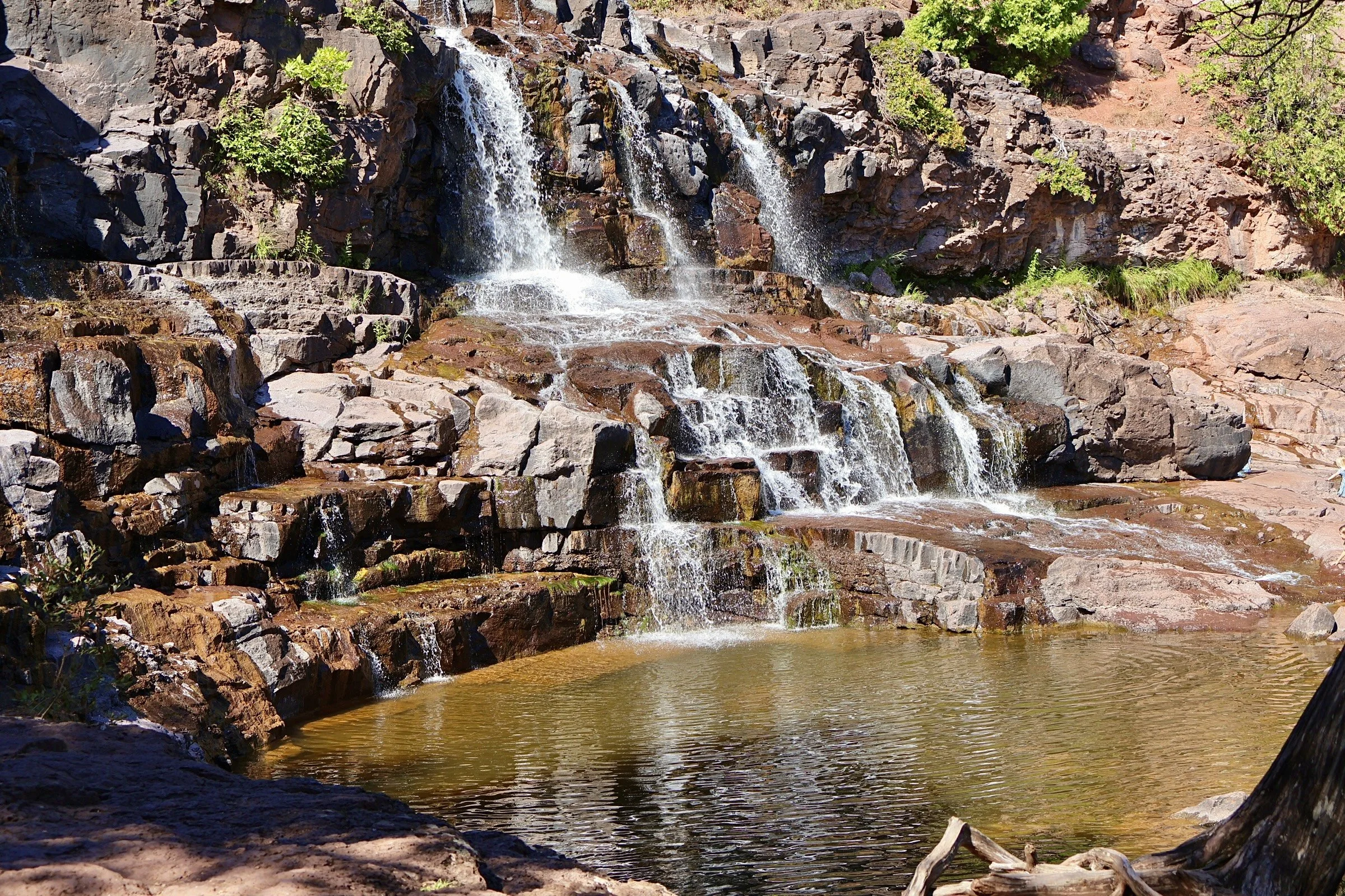 A small waterfall cascading over rocks in a natural outdoor setting with green foliage and a pool of water at the bottom.