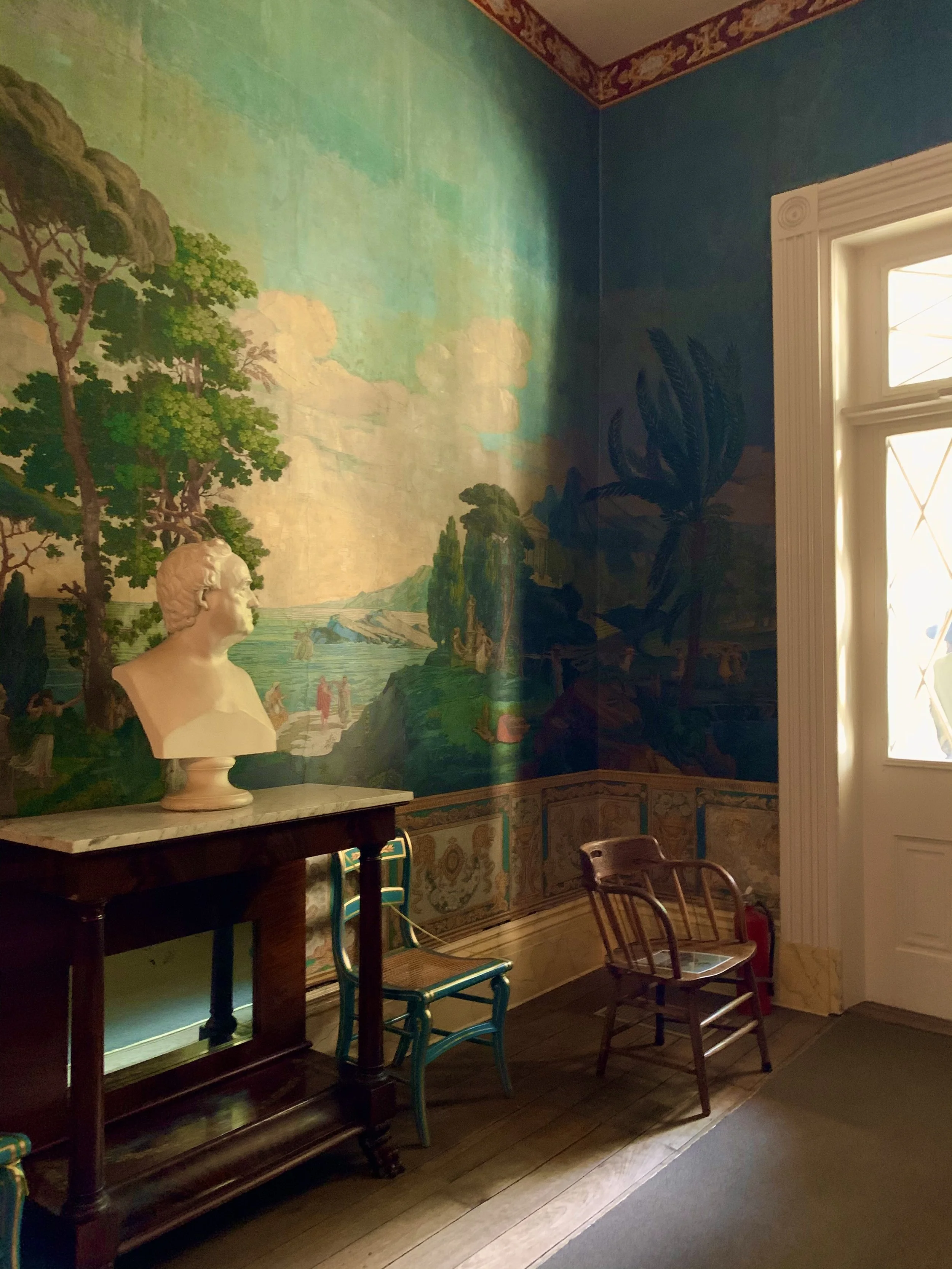A wall mural of a landscape with trees, water, and sky, with a bust sculpture on a table in front, in a room with chairs and window.
