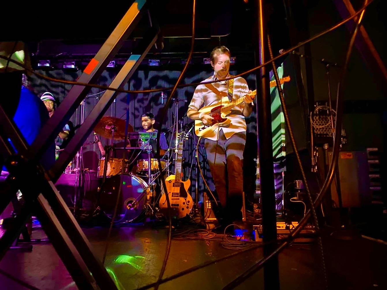 Band performing on stage, with a guitarist in the foreground, drummer in the background, and colorful stage lighting.