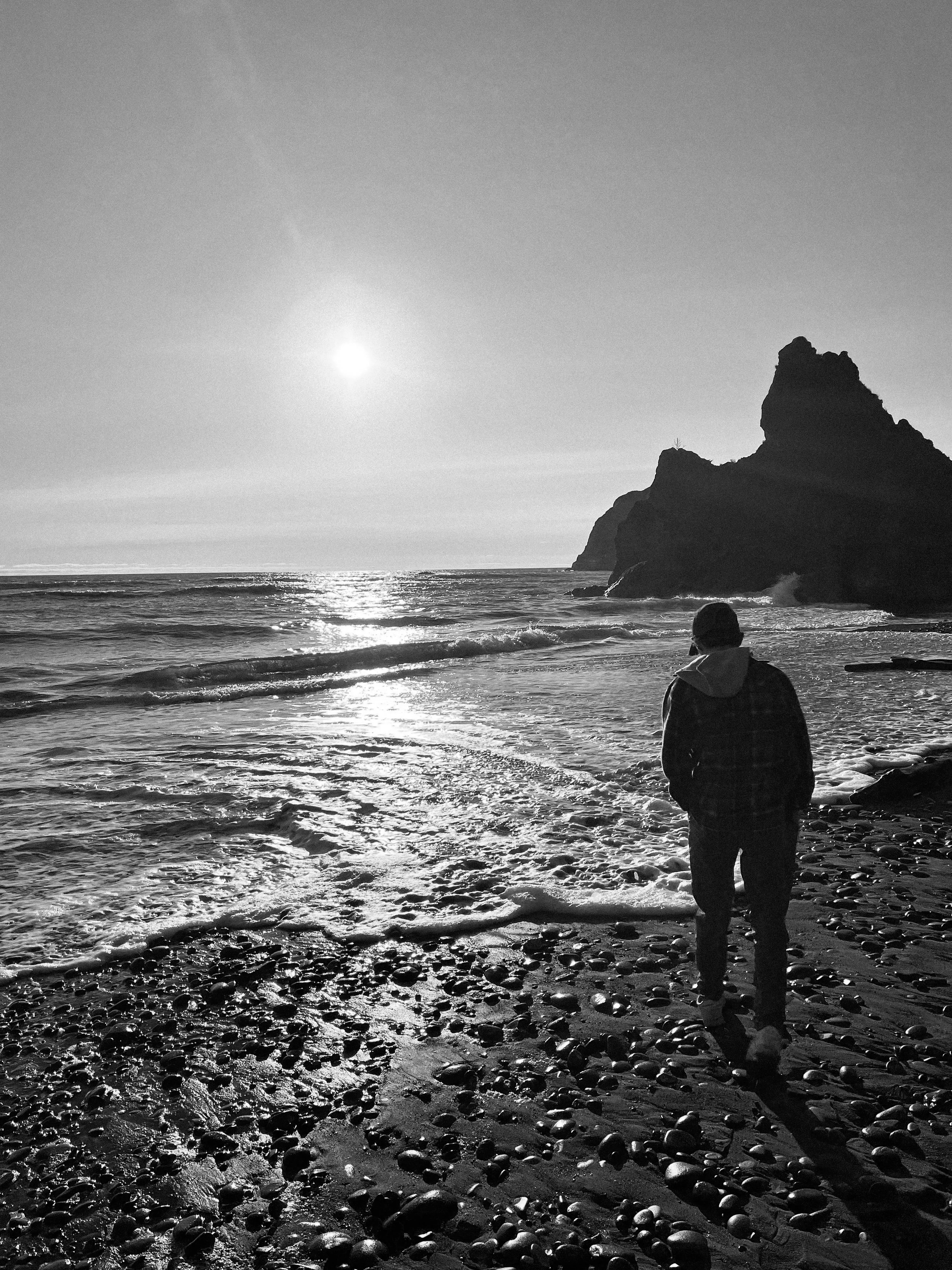 A person walking on a rocky beach with a large rock formation in the distance and the sun setting or rising in the sky, creating reflections on the water.