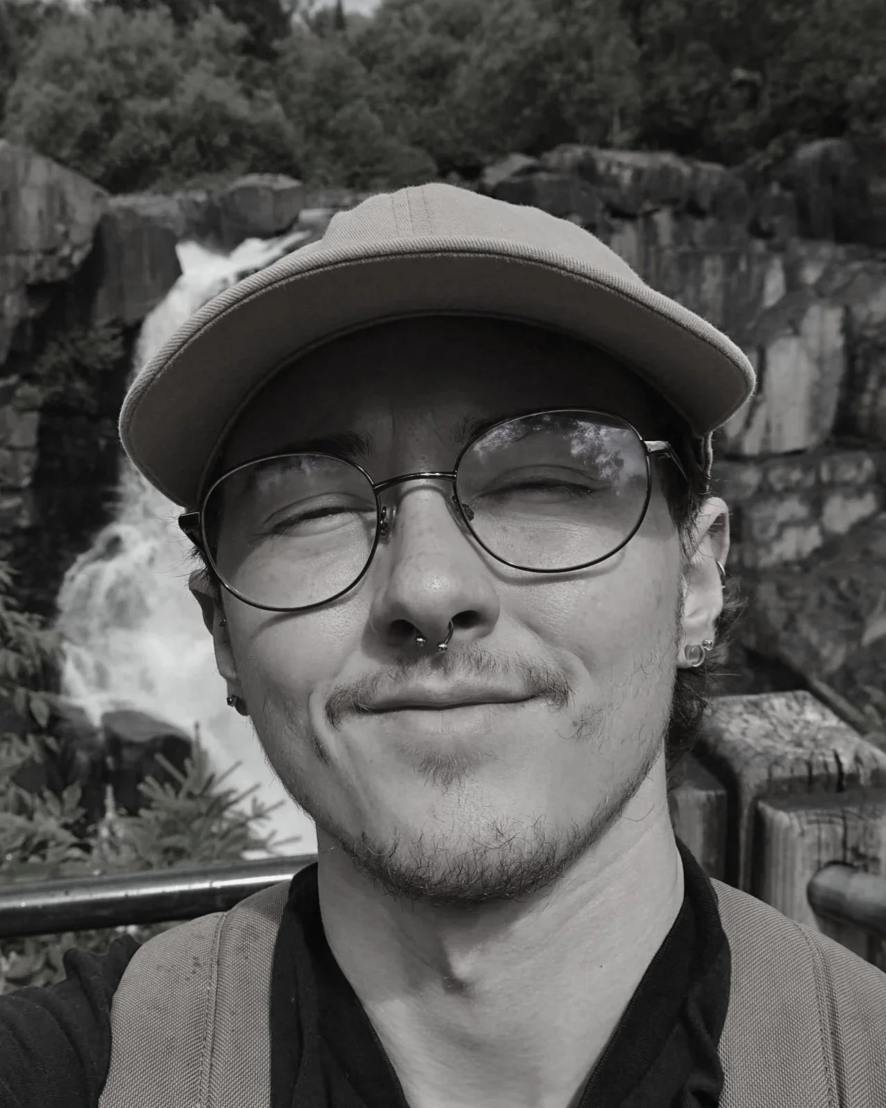 A person with glasses, a cap, and facial piercings smiling outdoors in front of a waterfall and rocky landscape.