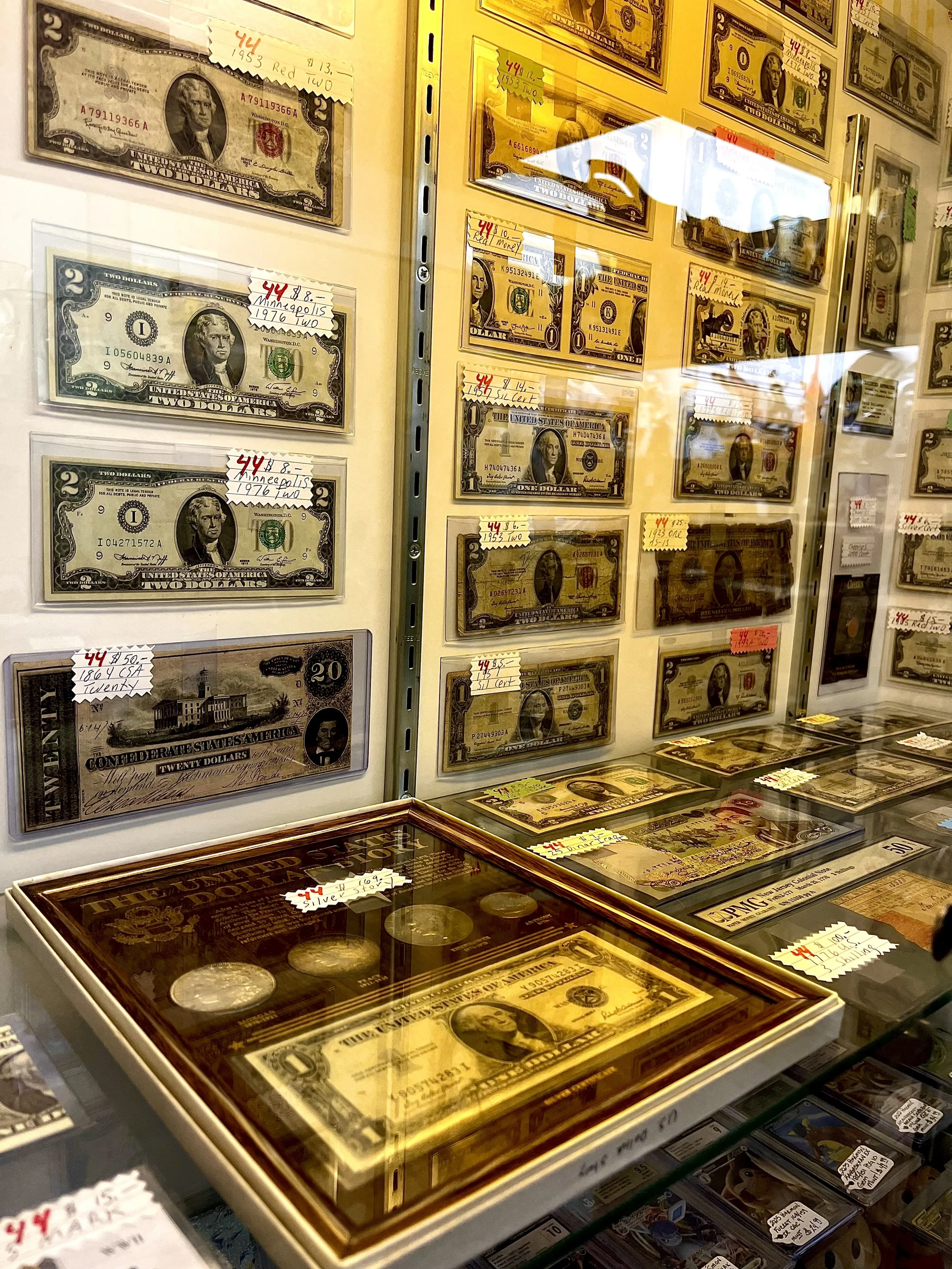 Display case with various collectible currency notes, mostly U.S. dollar bills, with some coins in a glass case and small handwritten price tags attached to each item.