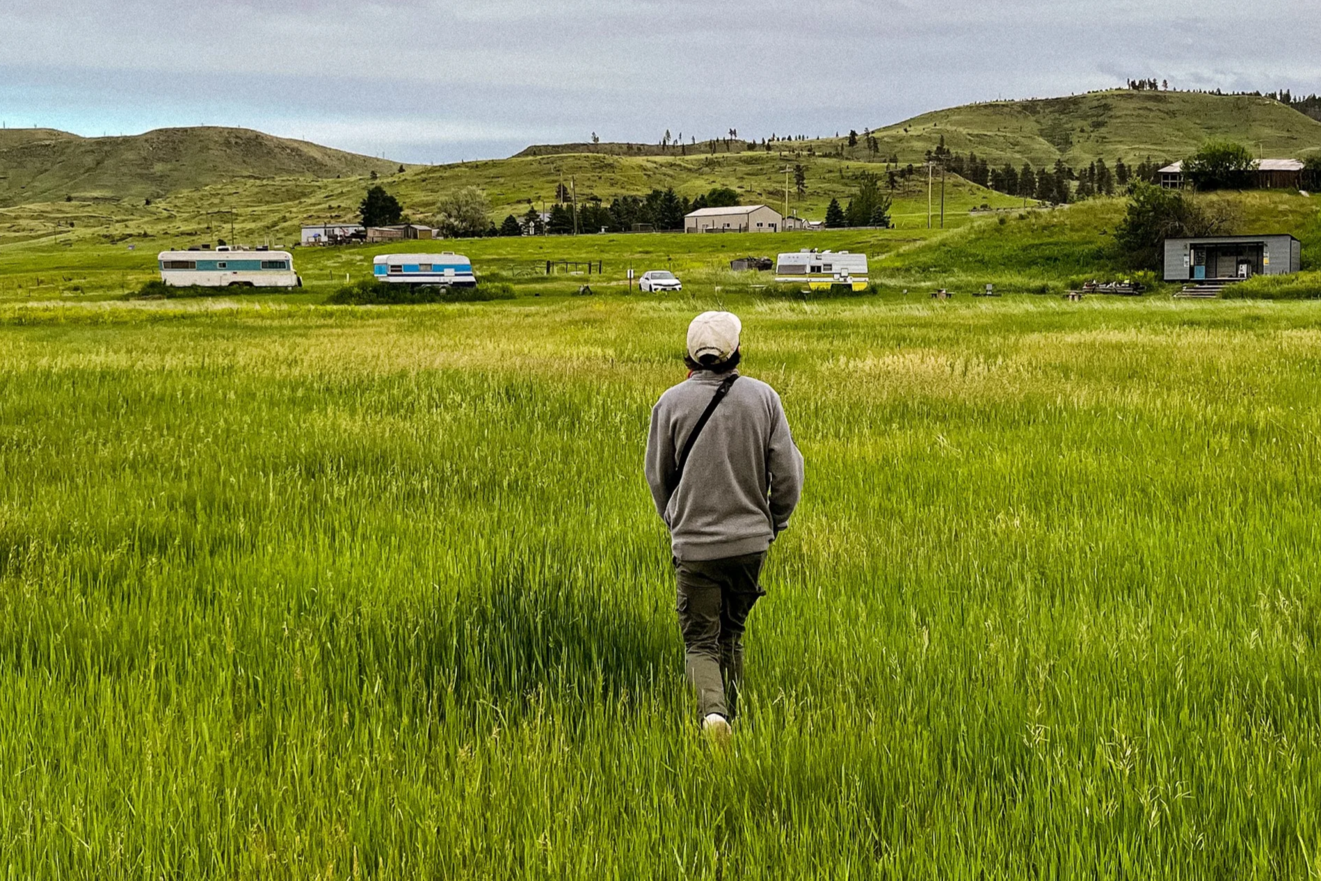 A person walking through a green field with hills and RVs in the background under a cloudy sky.