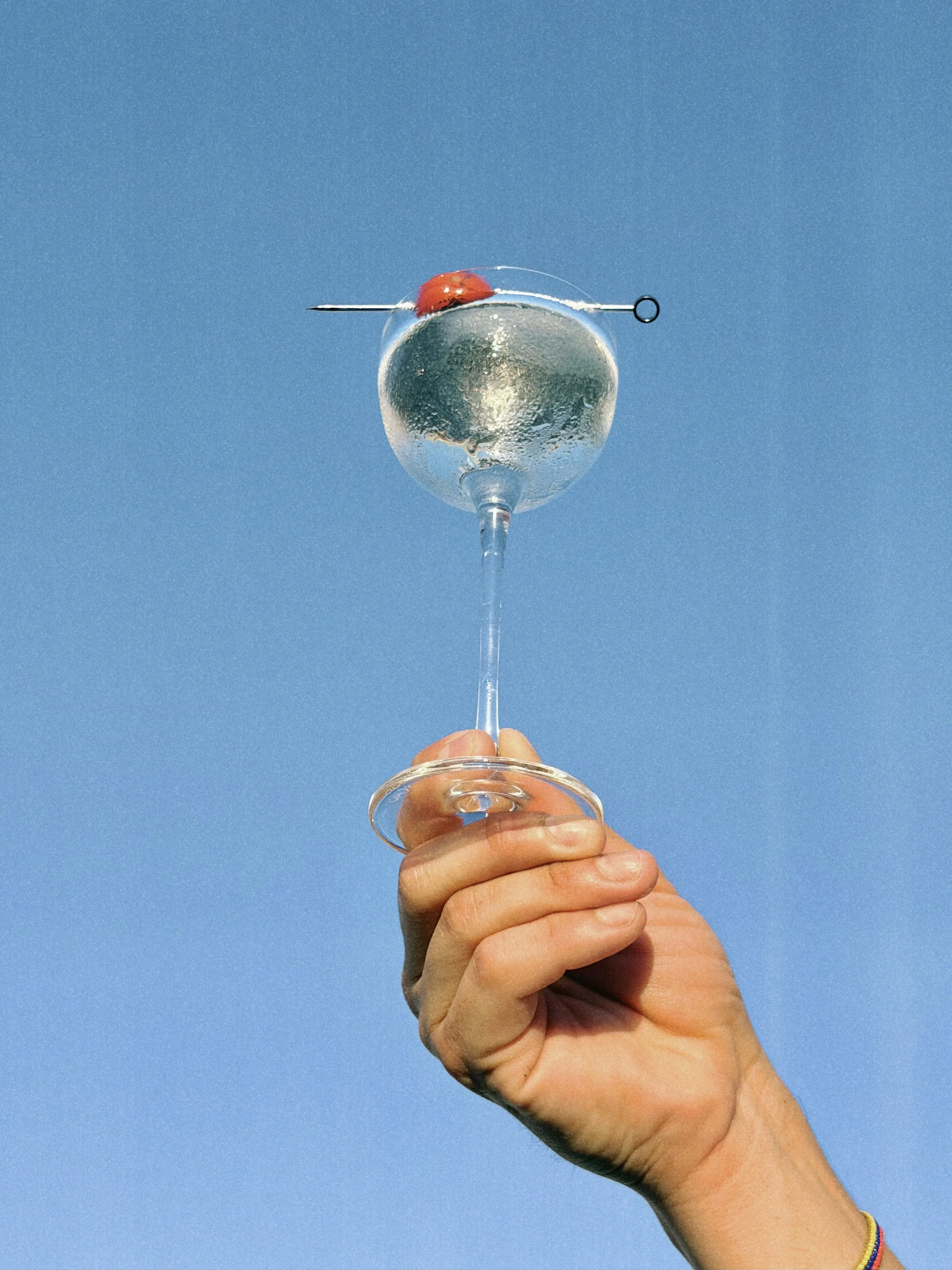 Hand holding a glass of water against a clear blue sky, with a red and black ladybug on the rim of the glass.