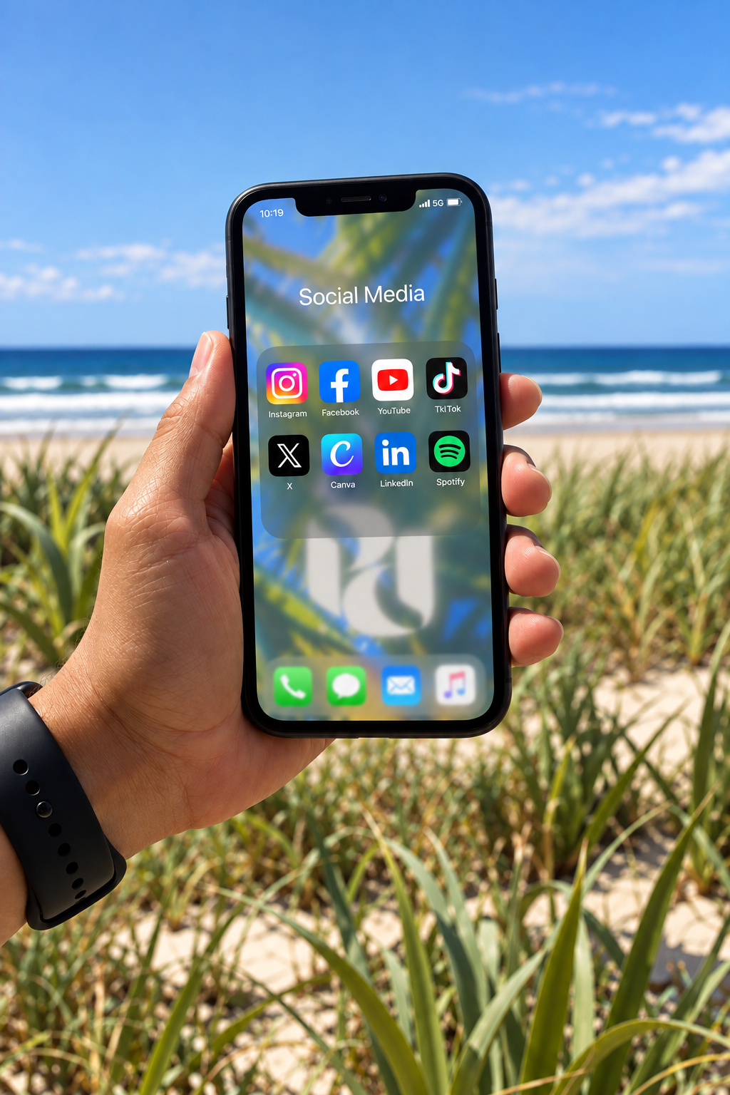 A person holding a smartphone in front of a beach with grassy dunes and the ocean in the background. The phone screen displays a folder labeled 'Social Media' with icons for Instagram, Facebook, YouTube, TikTok, X, Canva, LinkedIn, and Spotify.