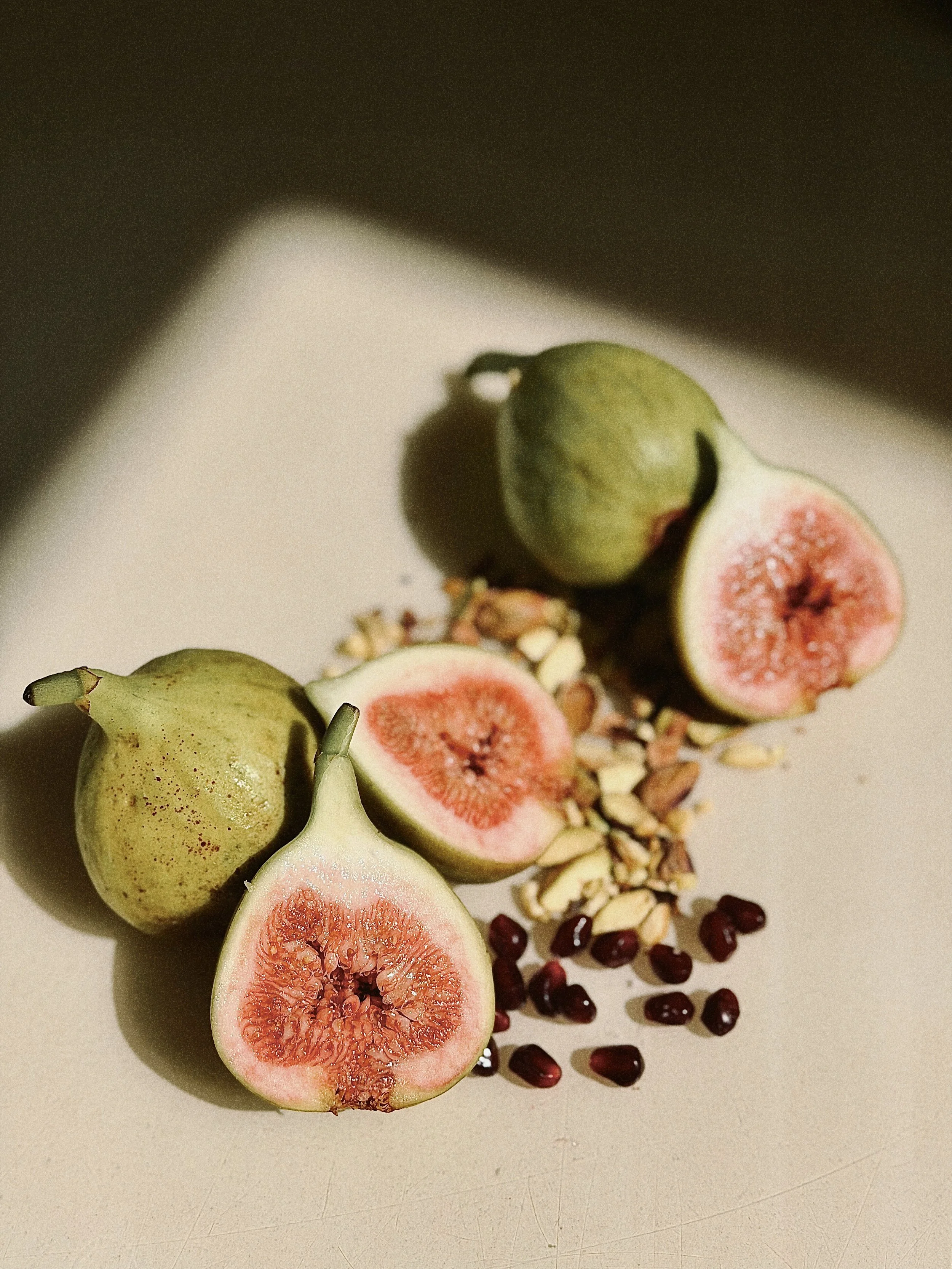 Halved fresh figs, whole figs, pomegranate seeds, chopped nuts, and other seeds arranged on a white surface.