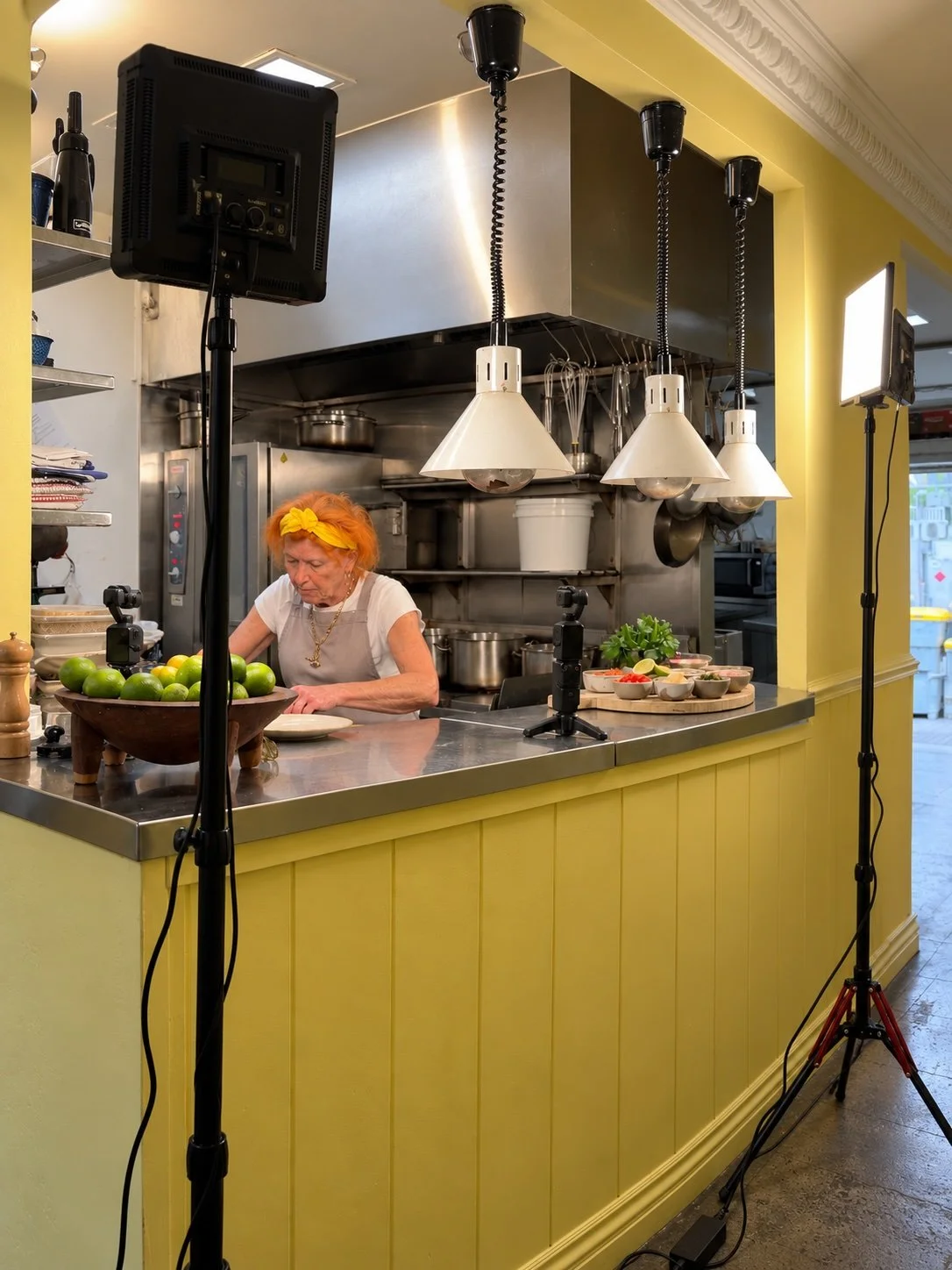 A woman with orange hair and a yellow headscarf working in a kitchen behind a yellow counter, preparing food with various ingredients and kitchen utensils, with professional lighting and filming equipment around her.