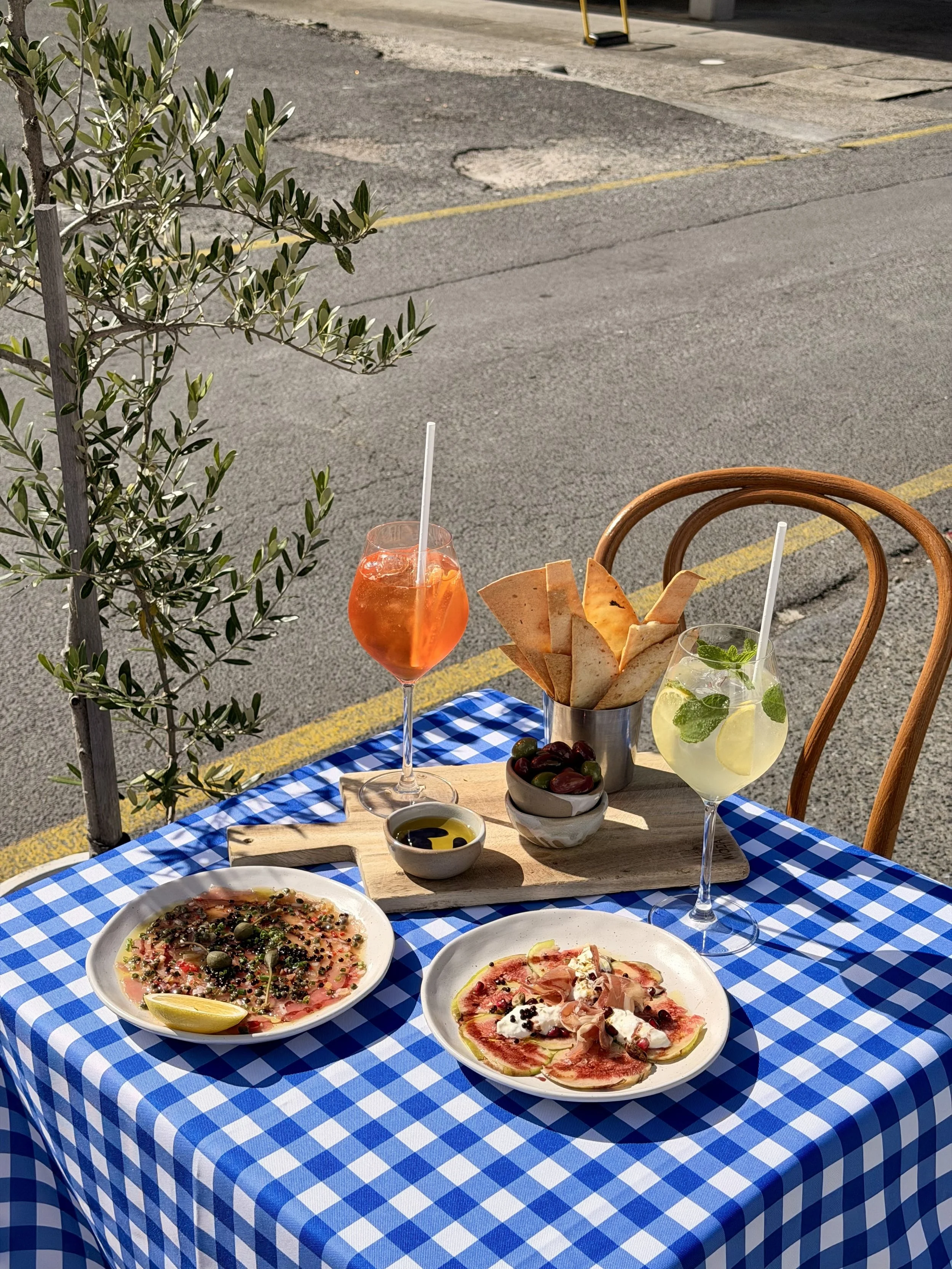 Outdoor restaurant table with blue and white checkered tablecloth, featuring three pizzas on white plates, a wooden serving board, a bowl of olives, and two colorful cocktails, one orange and one lemon-mint, on a sunny day.