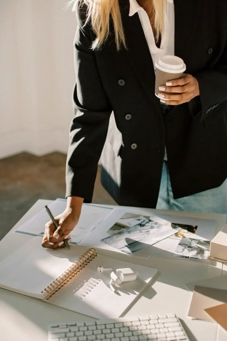 A woman in a black blazer and white shirt writing on a large notepad at a desk, holding a paper cup of coffee.