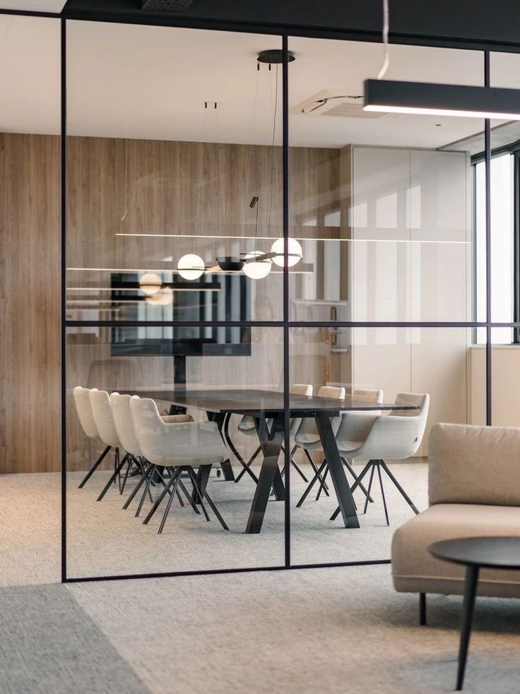 Modern conference room with a round table, eight beige chairs, a wall-mounted flat-screen TV, geometric black chandelier, glass walls, and a beige sofa in the foreground.